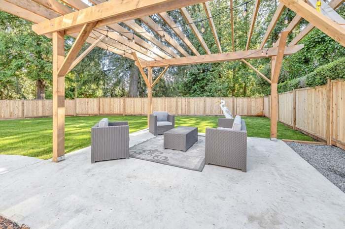 Grey wicker furniture under a wooden pergola with string lights in a fenced backyard.