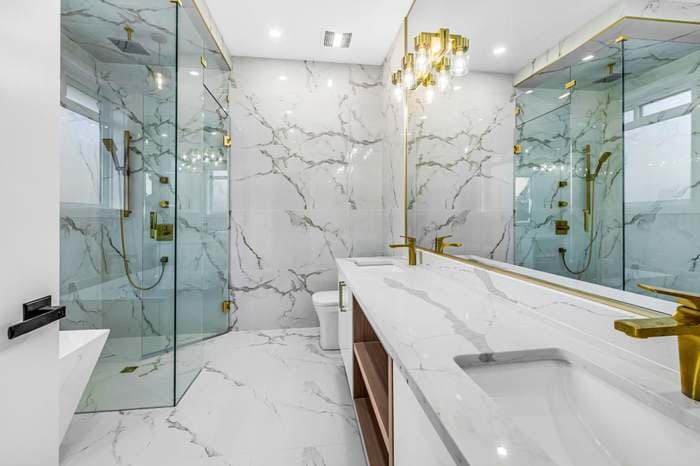 Luxurious white marble bathroom with gold fixtures, glass-enclosed shower, and a long double sink vanity.